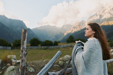 Fototapeta premium Young woman with long brown hair stands near a wooden fence in a mountain meadow, wrapped in a soft gray knit shawl, gazing into the distance with a peaceful, thoughtful mood.