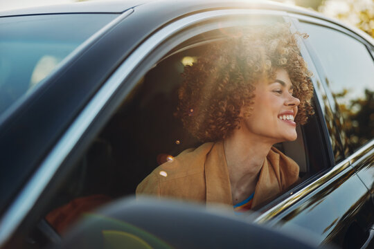 A joyful woman sits in a car with the window down, smiling as warm sunlight spills across her face and highlights her curly hair in a candid moment.