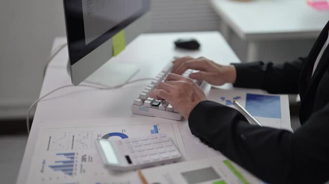 Business professional typing on computer keyboard with financial documents and tablet