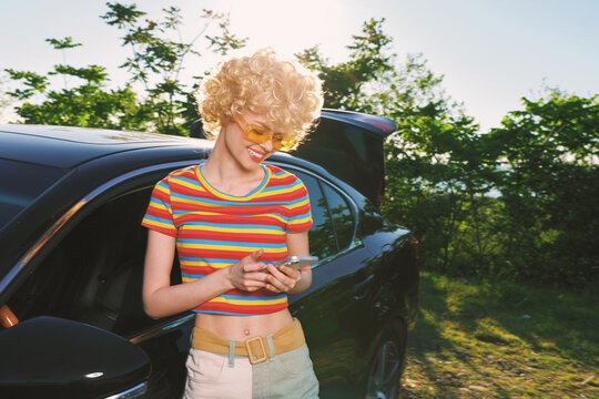A woman with a retro hairstyle in a striped crop top stands beside a car, looking at her phone in a sunlit outdoor setting. Concept for casual style and modern connectivity.