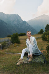 Fototapeta premium Smiling woman in cozy sweater sitting on rock in mountain meadow, autumn landscape, casual outfit, relaxed mood, natural light, outdoor lifestyle