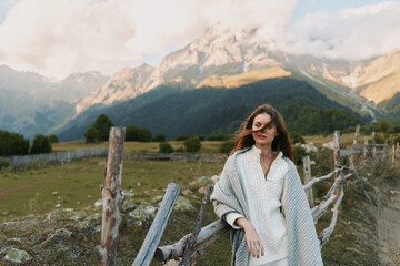 Fototapeta premium A young woman in a cozy knit poncho stands beside a wooden fence in a mountain valley, gazing ahead with a calm, reflective mood.