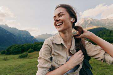 Fototapeta premium A joyful woman outdoors, wearing a light jacket, backpack over her shoulder, smiling as green fields and distant mountains frame the scene. Lifestyle moment of freedom and nature.