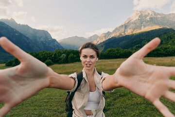 Fototapeta premium Young woman with a backpack reaches out towards the camera while hiking in a mountainous landscape under a partly cloudy sky.