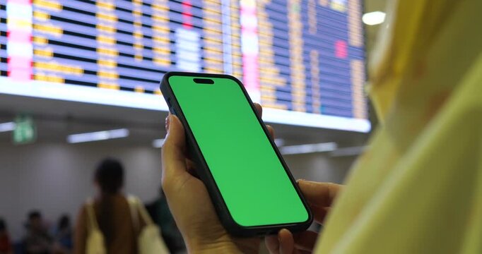 Cinematic close-up of traveler hands holding smartphone with green screen chroma key mockup inside airport terminal, blurred flight board background