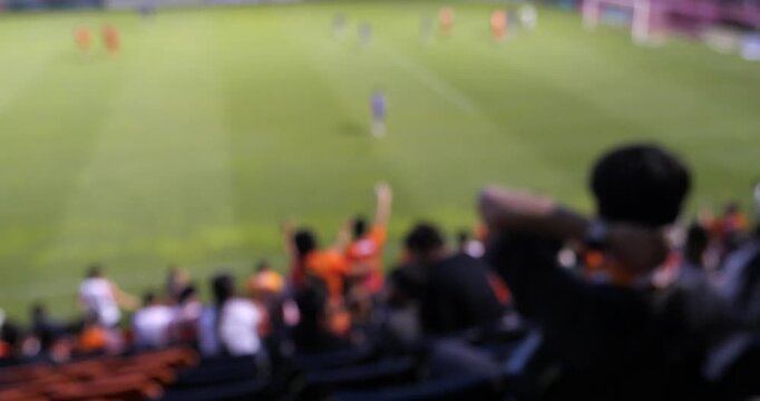 Cinematic wide shot of football fans silhouette cheering on stadium grandstand seats, out of focus blurred green soccer pitch background