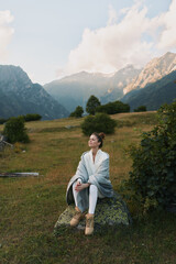Fototapeta premium A young woman sits on a rock in a meadow with distant mountains, wearing a light cardigan and boots, looking calm and thoughtful on a sunny day.