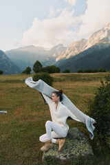 Fototapeta premium A woman in a white sweater and boots leaps across a meadow, with mountains in the background, capturing a vibrant outdoor lifestyle moment in nature.