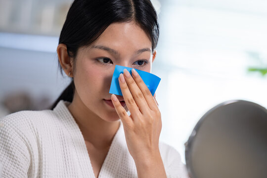 Focused serious asian woman looking at round mirror using blue blotting paper to remove excess oil from face during morning skincare beauty routine in bathroom