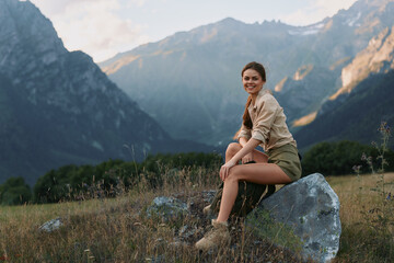 Fototapeta premium A young woman sits on a rock in a mountain meadow, smiling softly as she enjoys a peaceful, outdoor escape with rugged peaks in the distance.