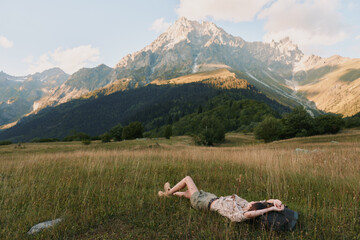 Fototapeta premium A woman in casual clothes lies on grass in a mountain meadow, relaxing under a partly cloudy sky with tall peaks in the background, enjoying nature and lifestyle outdoors.