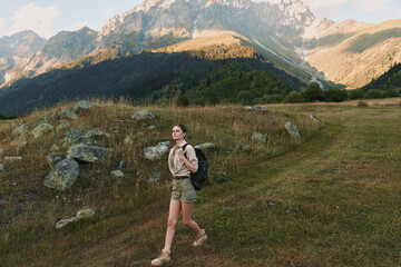 Fototapeta premium Woman hiking through a mountain meadow with a backpack, exploring nature on a dirt trail, showcasing an active outdoors lifestyle, calm adventurous mood in a wide open alpine landscape