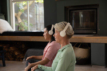 Fototapeta premium Two mature women sitting on floor in home wellness area, wearing over-ear headphones, facing bench
