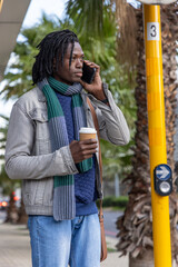 Fototapeta premium Mid-adult African American man wearing scarf standing near palms holding phone and coffee by pole