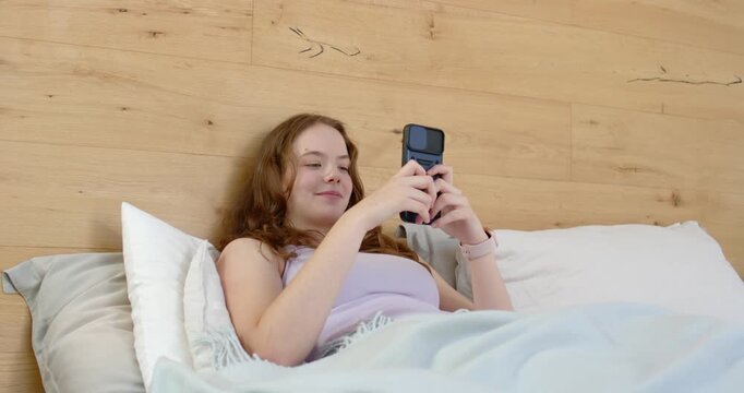 Adult woman reclining on bed in lilac tank top holding flip smartphone, reading message, smiling