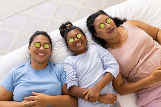 Diverse female family with youth lying on white bedding at home in loungewear with cucumber slices