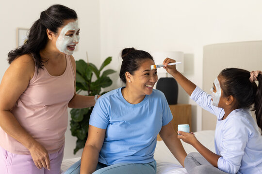 Diverse female family sharing skin care in bedroom, teenage girl applying mask with cream jar