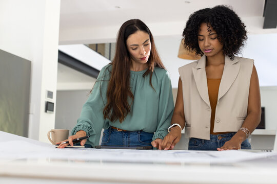 Two female colleagues leaning over large blueprints on table in bright studio, marking on tablet