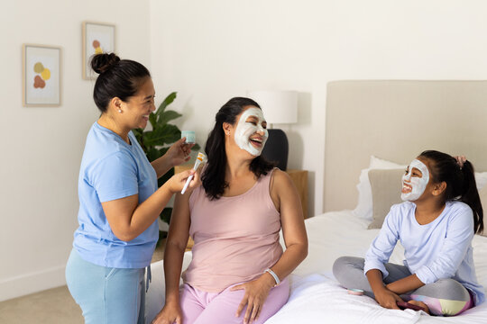 Diverse female family on bed in softly decorated bedroom, applying masks with jar and facial brush