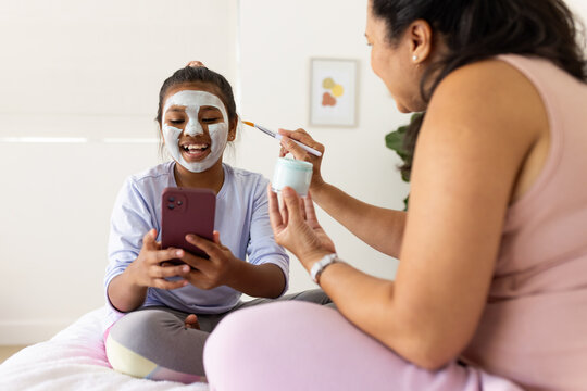 African American mother and youth daughter sitting on bed, laughing holding smartphone and mask jar