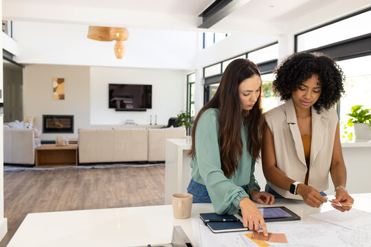 Diverse female team standing, reviewing tablet, plans at kitchen island, holding swatch, copy space