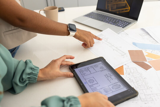 Female colleagues pointing and interacting with tablet at work table with plans, swatches, laptop