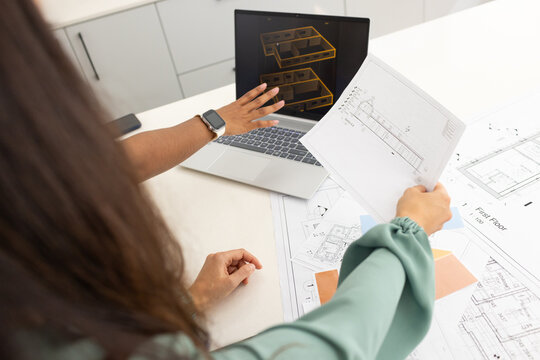 Diverse female coworkers reviewing blueprints on white table, pointing at laptop 3D wireframe
