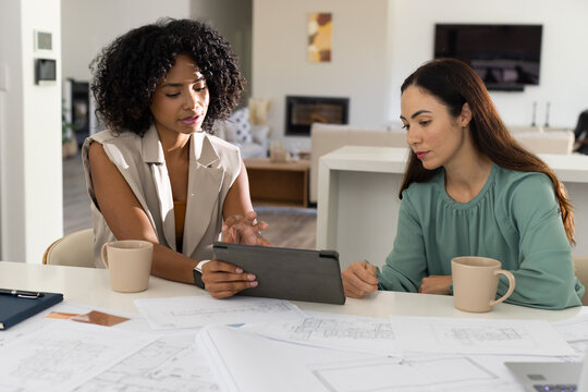 Diverse female colleagues reviewing tablet and blueprints in open-plan living room with coffee mugs