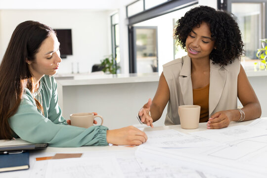 Diverse female designers reviewing design plans at modern office, pointing at drawings holding mugs