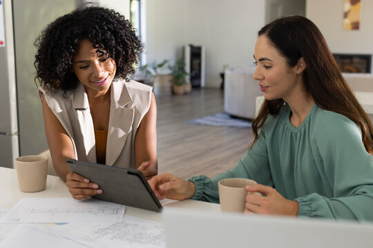 Diverse female colleagues in beige vest and teal blouse reviewing printed plans and tablet at home