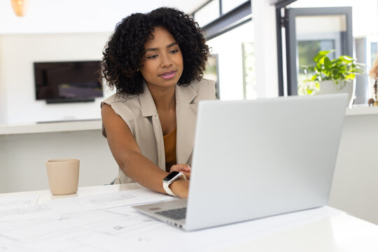 Adult woman wearing sleeveless blazer working on laptop with printed plans in open-plan workspace