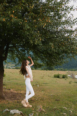 Fototapeta premium Cheerful woman outdoors, standing in a sunlit field under a leafy tree, reaching up with relaxed pose, white outfit, natural colors, carefree mood
