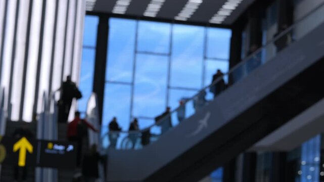 People move on escalators inside a modern transit station. Blurred background shows large windows and structural beams. Natural light streams through glass walls