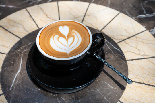 A beautifully crafted latte art on a black ceramic cup, placed on a marble saucer with intricate designs. The art features a heart shape, symbolizing love and care, set against a rich, creamy