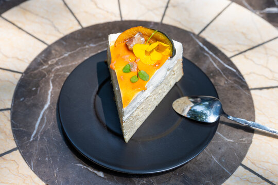 A slice of vibrant yellow and orange cheesecake sits on a black marble plate, surrounded by a luxurious marble setting. The cheesecake, topped with a golden-brown fruit compote and green pistachio
