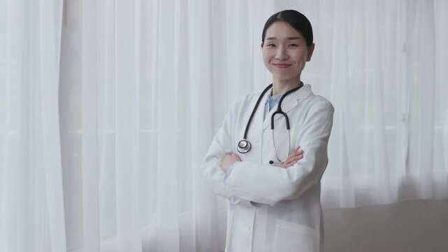 Asian Female Doctor in White Coat Smiling with Arms Crossed by Bright Window
