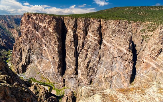 View of the massive Painted Wall in Black Canyon of the Gunnison National Park. White pegmatite dikes create stripes on the dark, sheer cliff. The Gunnison River is visible below