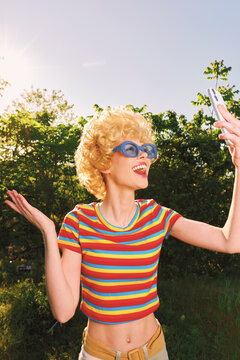 Young woman with curly blonde hair wearing a striped rainbow shirt and blue sunglasses poses for a joyful outdoor selfie in a sunlit park, the bright colors echo a retro vibe and a carefree outdoor