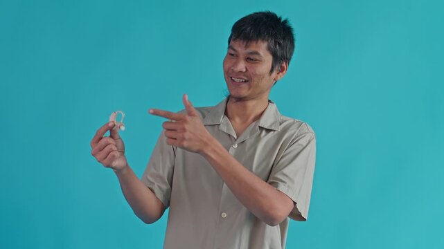 Happy Young Man Presenting And Pointing To Modern Hearing Aid Device For Auditory Health And Hearing Loss Solution On Blue Backdrop