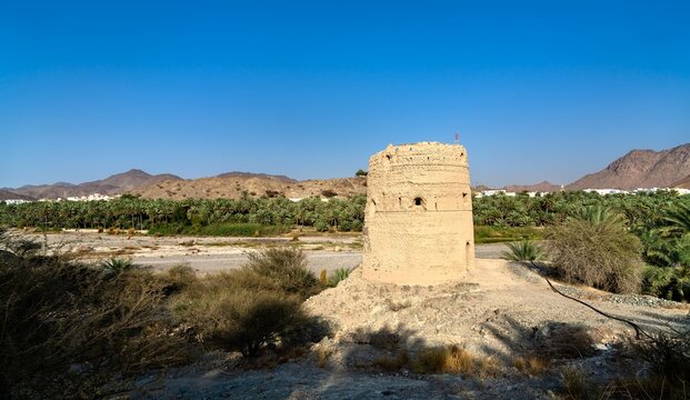 The historic stone structure of Al Basra Fort Tower stands near a lush palm oasis and dry riverbed in the scenic mountain landscape of Fanja, Oman.