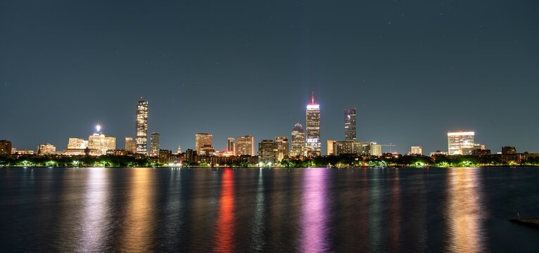 Boston, United States, glows across the Charles River on a clear night, its towers casting colorful reflections onto calm water when seen from Cambridge's esplanade.