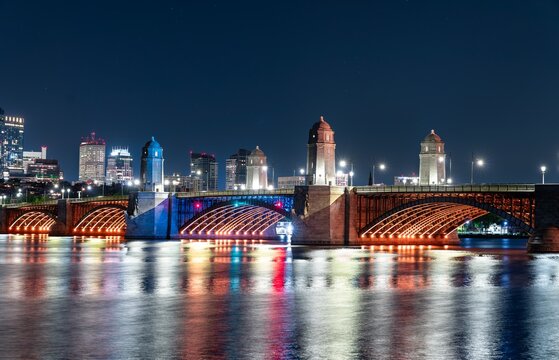 Night view of the Longfellow Bridge in Boston glowing with orange lighting, arching over the Charles River with reflections shimmering on the water and the city skyline in the background
