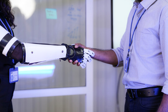 An AI software architect testing human-robot interaction by shaking hands with a cybernetic bionic prosthetic arm during a technology demo.