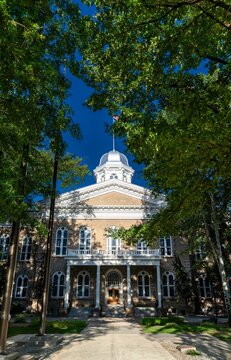 Vertical view of the historic Nevada State Capitol building in Carson City. The facade and silver dome are framed by lush green trees under a clear blue sky. Represents state government and history