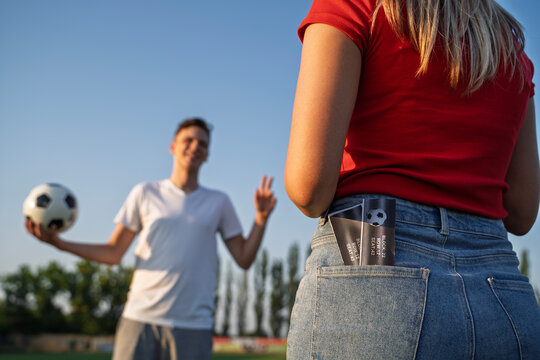 Back view, girl holding two soccer tickets in her back pockets of jeans, her fiend happy to see her in the back blurred 