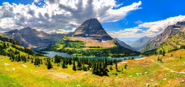 A wide panoramic view of Hidden Lake and Bearhat Mountain from the Logan Pass overlook in Glacier National Park, Montana. A green meadow is in the foreground under a dramatic cloudy sky