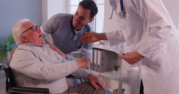 Male doctor in white uniform shaking hands with senior patient man with disability in wheelchair standing with nurse after medical exam in clinic. Medicine concept. 4k video.