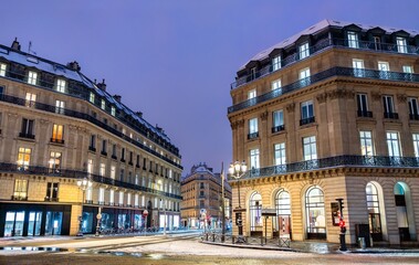 Fototapeta premium Snowy winter view of a street intersection and classic Haussmann buildings in Paris, France. The urban evening scene features snow on the road and glowing windows.