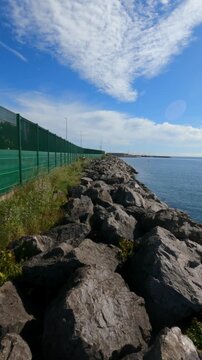 Static ground-level shot of rocky breakwater with tall green windbreak fence along Playa Arbeyal coastline. Calm blue sea and sky.