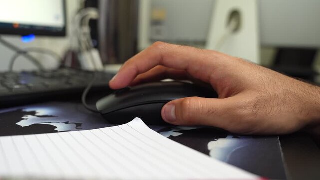 Close-up of a man's hand navigating with a black computer mouse on a desk in a home office setting. Features a keyboard and monitor in soft focus, representing productivity and digital work.